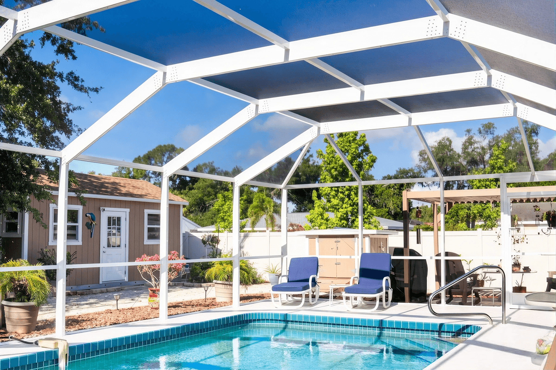 Sunny backyard swimming pool beneath a screened patio with blue shade panels, lounge chairs, tropical plants, and a calm, inviting atmosphere.