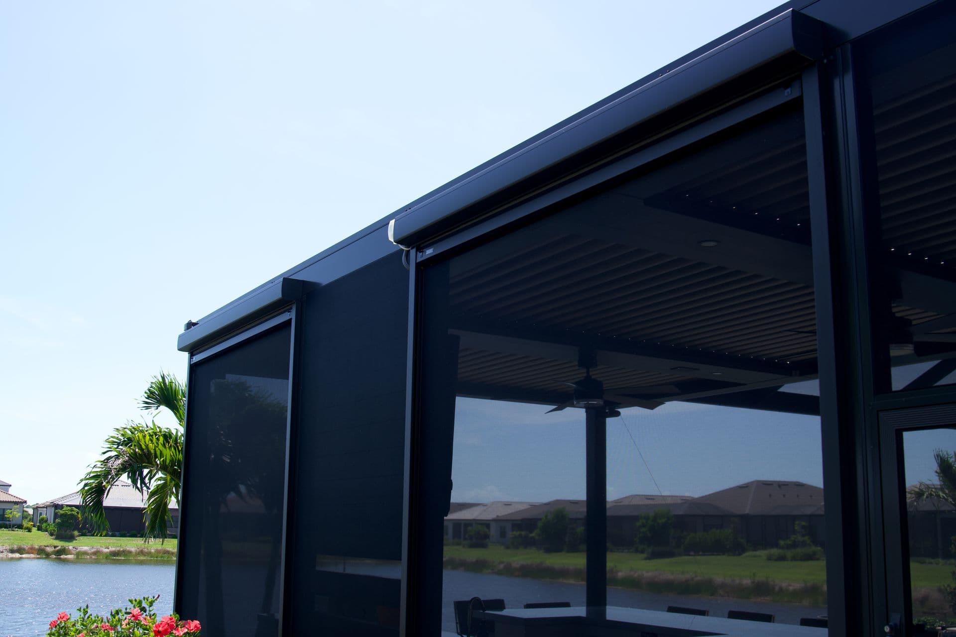 Modern black screened patio enclosure beside a pond, with palm trees, flowers, and homes beyond, creating a calm, sunny outdoor retreat.